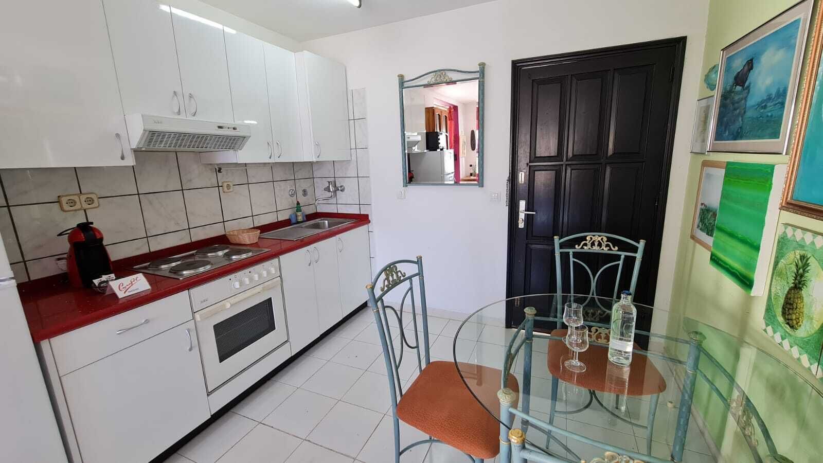 Una cocina compacta con armarios blancos, encimera roja y una mesa de vidrio con sillas.