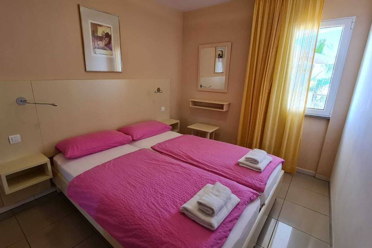 A double bed with pink bedding and two towels, a bedside table, a small shelf, and a window with yellow curtains.
