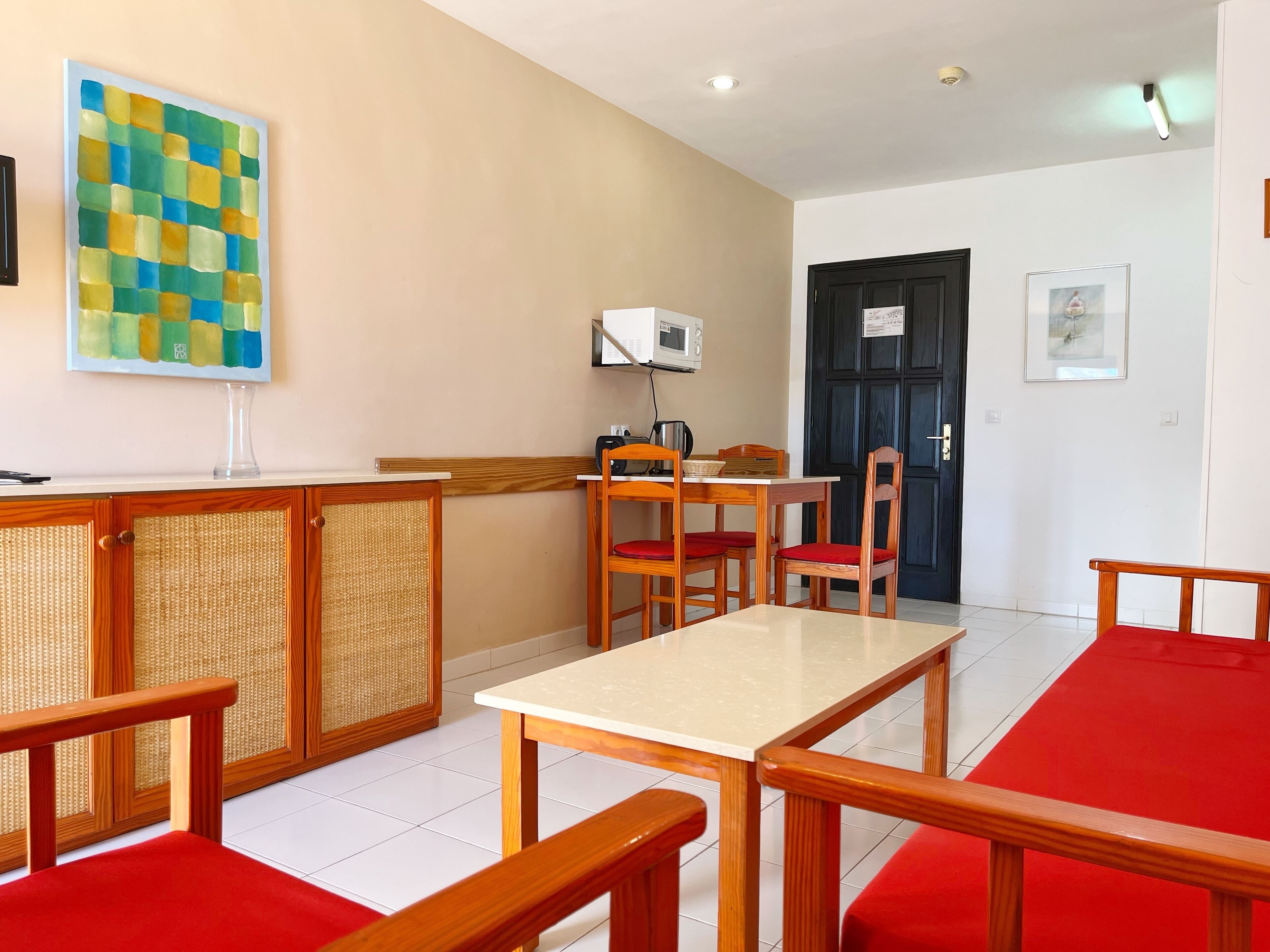 A hotel room with a wooden table and chairs, a small kitchenette, a door, and a wall-mounted TV.