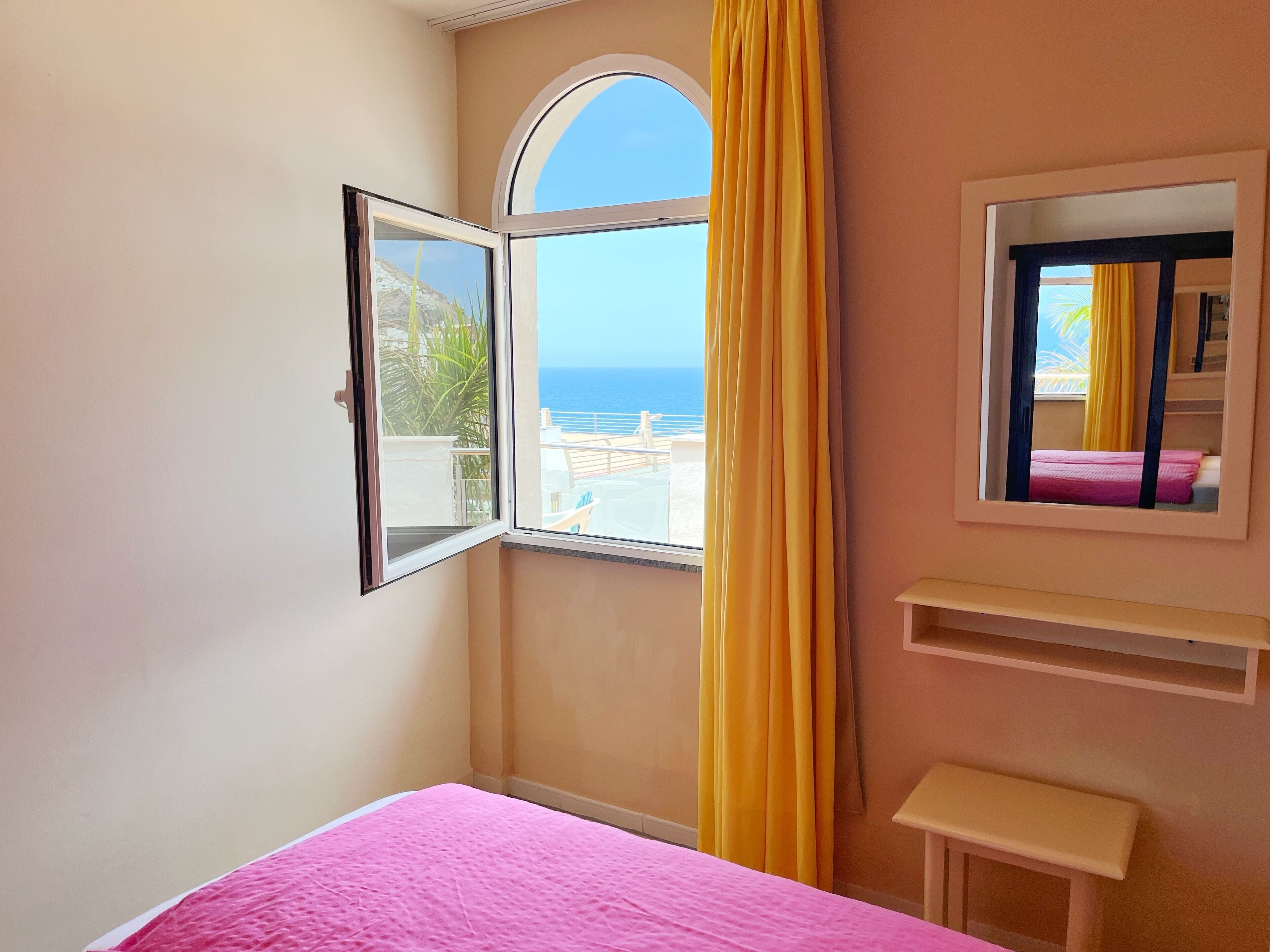 Um quarto com colcha rosa, janela com cortinas amarelas e vista para o mar.