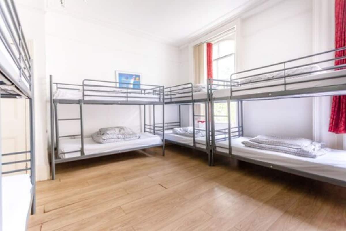 A dormitory room with three bunk beds, wooden flooring, and a window with red curtains.
