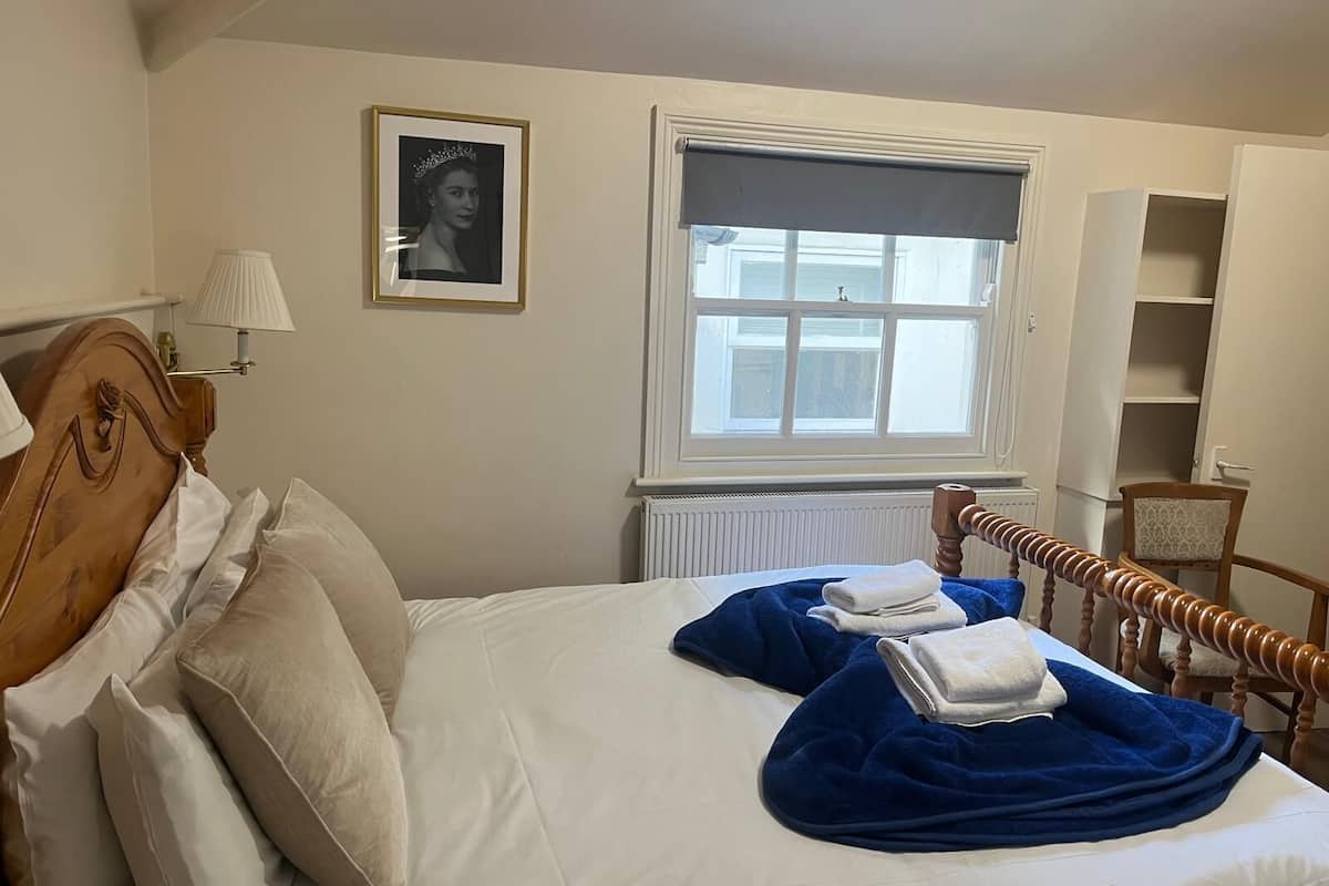 A neatly made bed with white and blue linens, a framed portrait on the wall, and a window with a white blind.