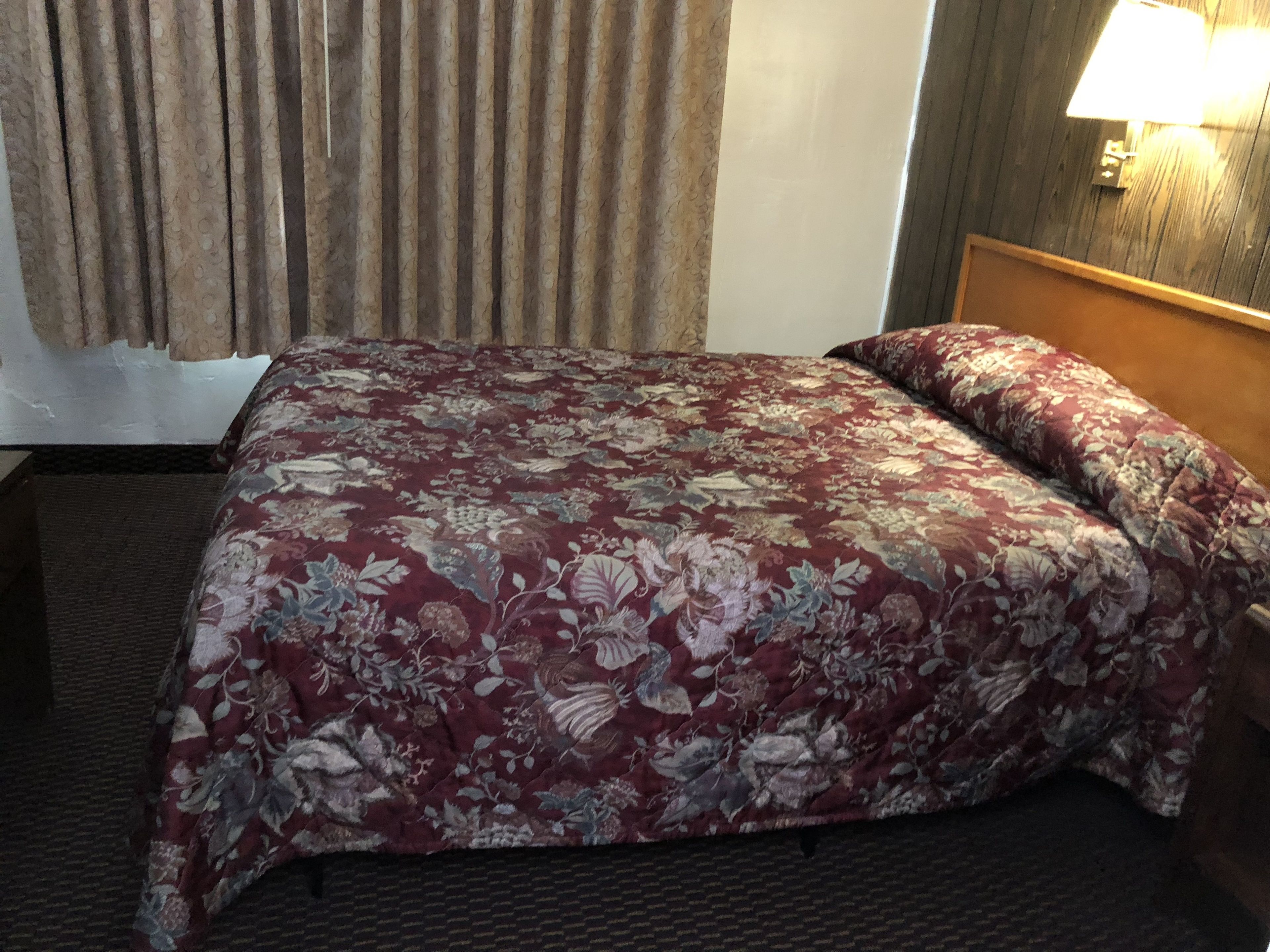 Habitación de hotel con cama, cabecero, lámpara de pared y ventana con cortinas.