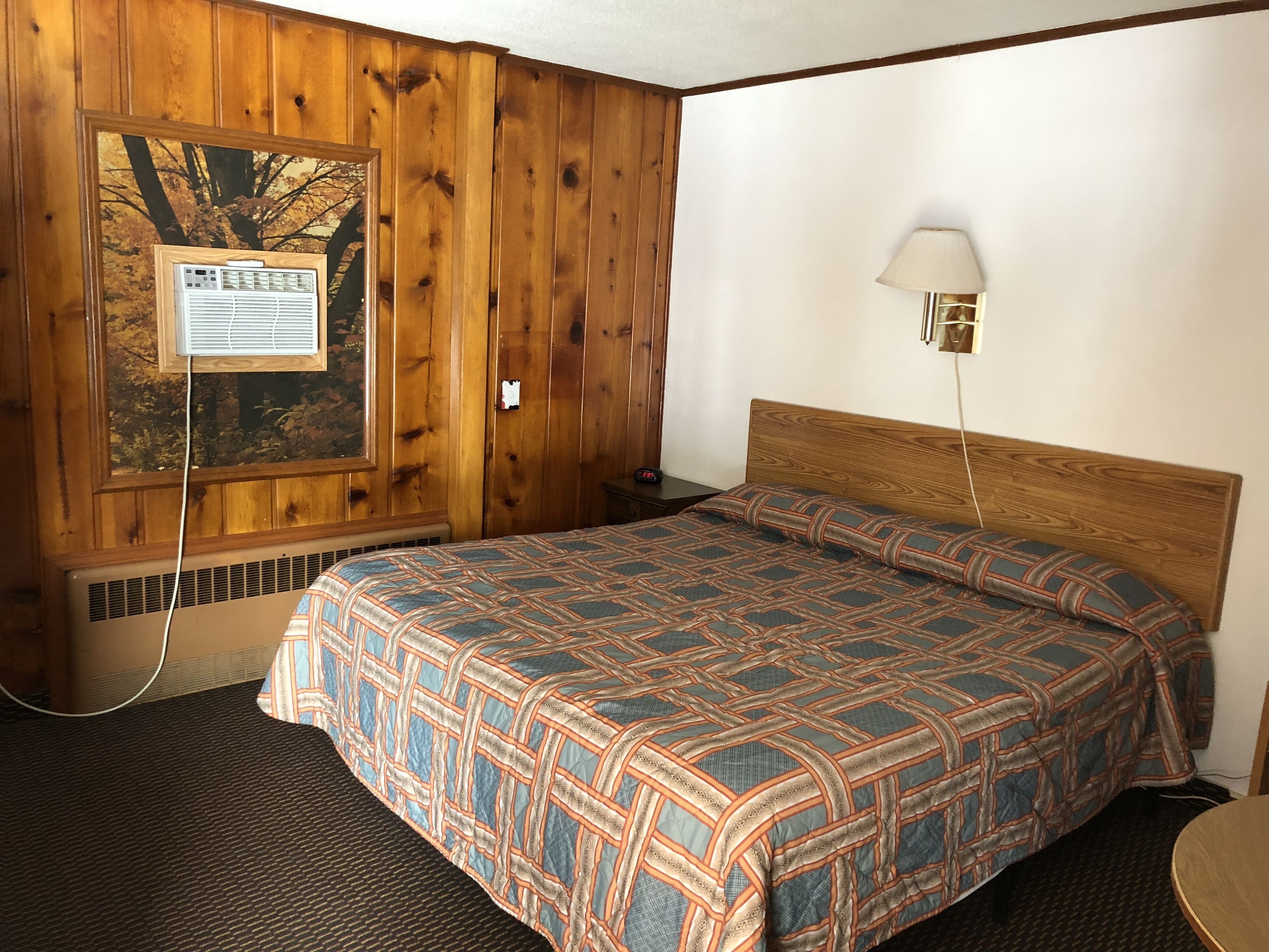Una habitación de hotel con un revestimiento de madera, una cama con colcha estampada, una mesita de noche con lámpara y un aviso en el marco en la pared.