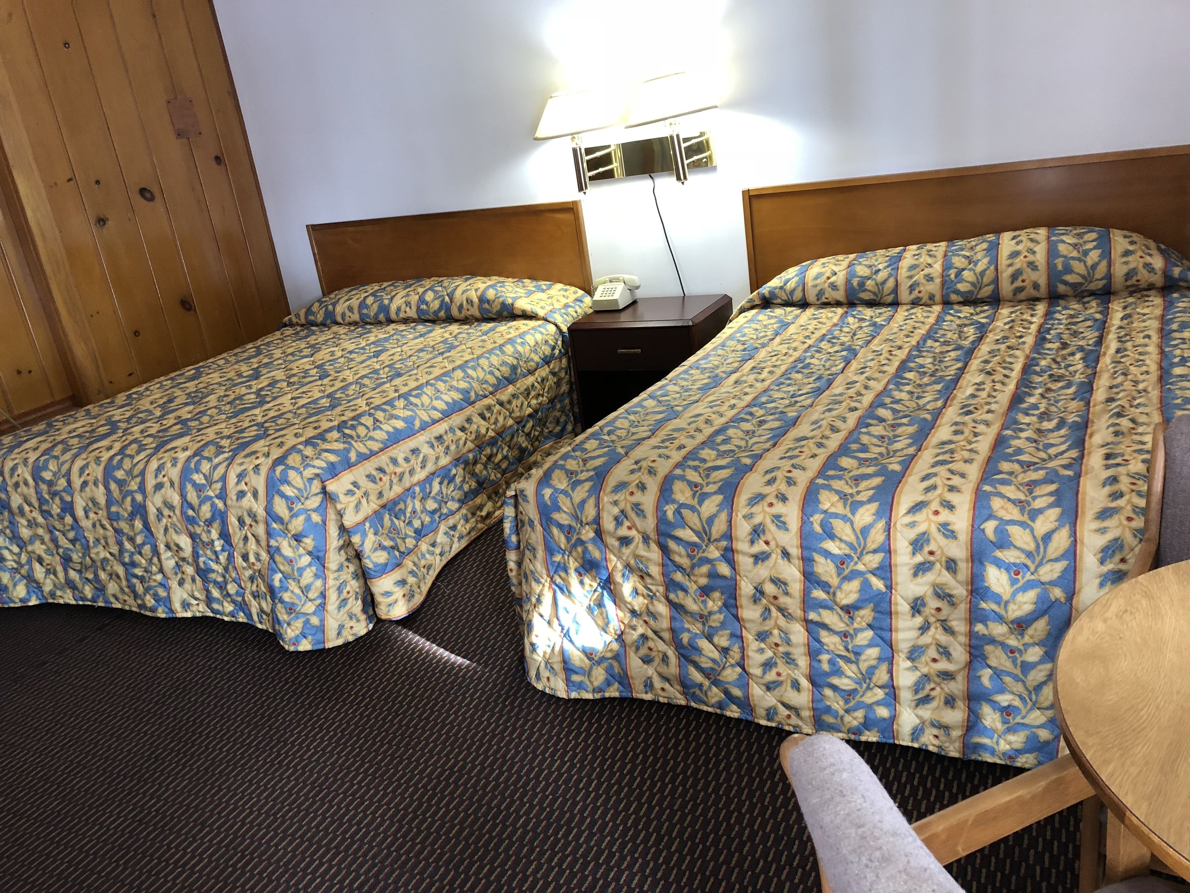 A hotel room with two single beds, a small wooden table, a nightstand with a lamp, and a wall-mounted air conditioning unit.