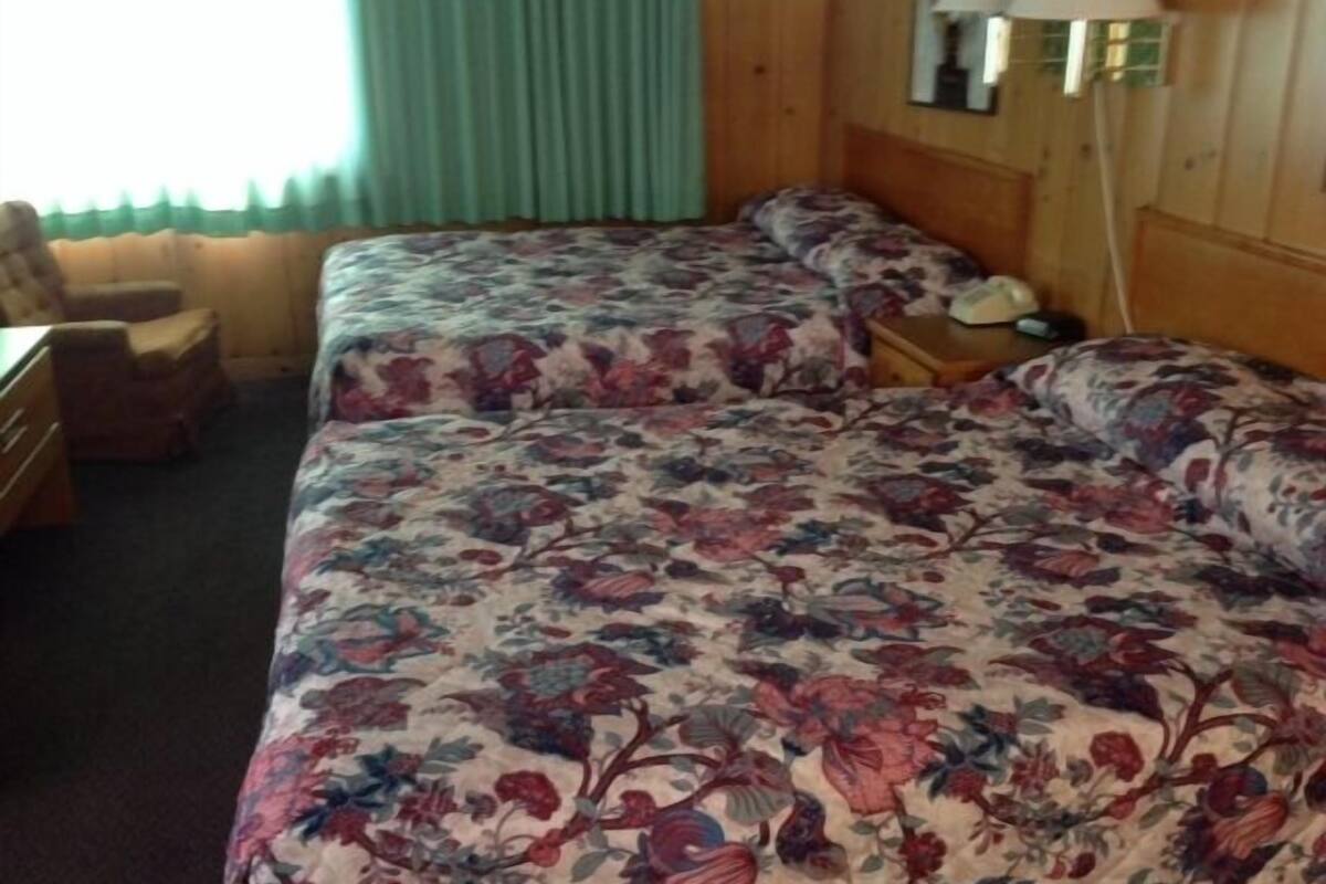 A hotel room with two beds, a wooden headboard, a desk, a chair, and a lamp.