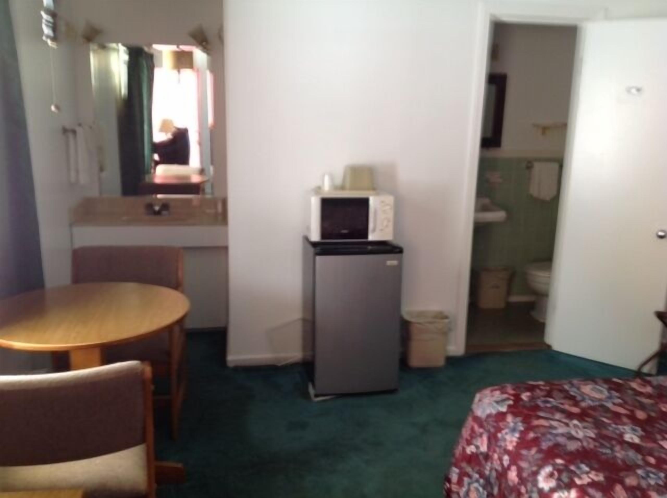 A compact hotel room with a bed, a small kitchenette with a refrigerator and microwave, a bathroom with a toilet and sink, and a dining area with a table and chairs.