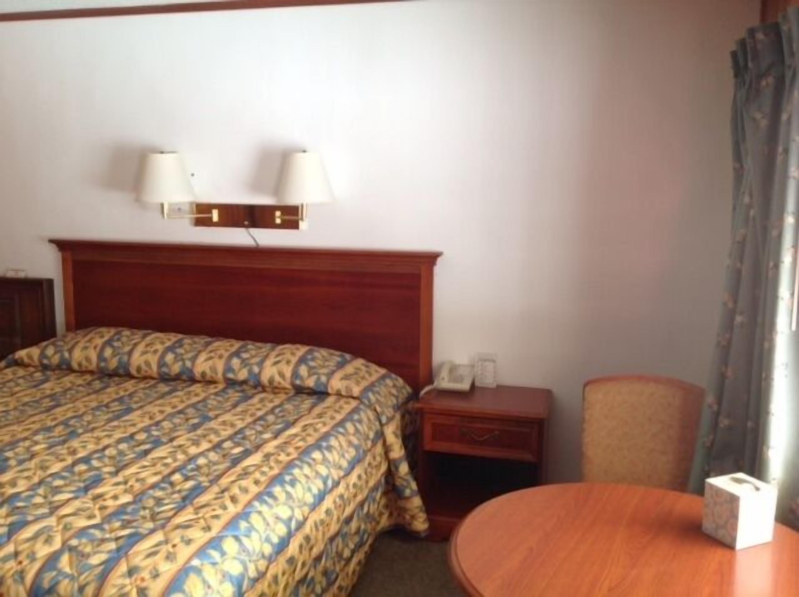 A hotel room with a large bed, a bedside table with a lamp, a chair, and a small table with a box.