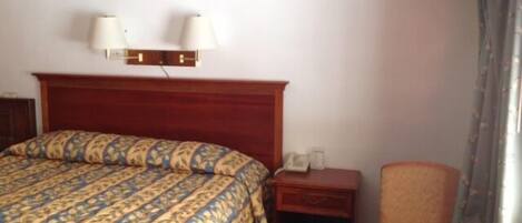 A hotel room with a large bed, a bedside table with a lamp, a chair, and a small table with a box.