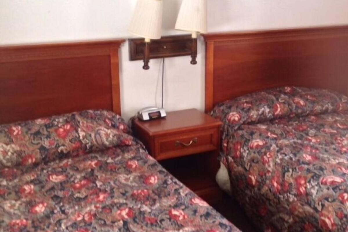 A room with two beds, each with floral bedding, a bedside table with a lamp, and a small drawer.