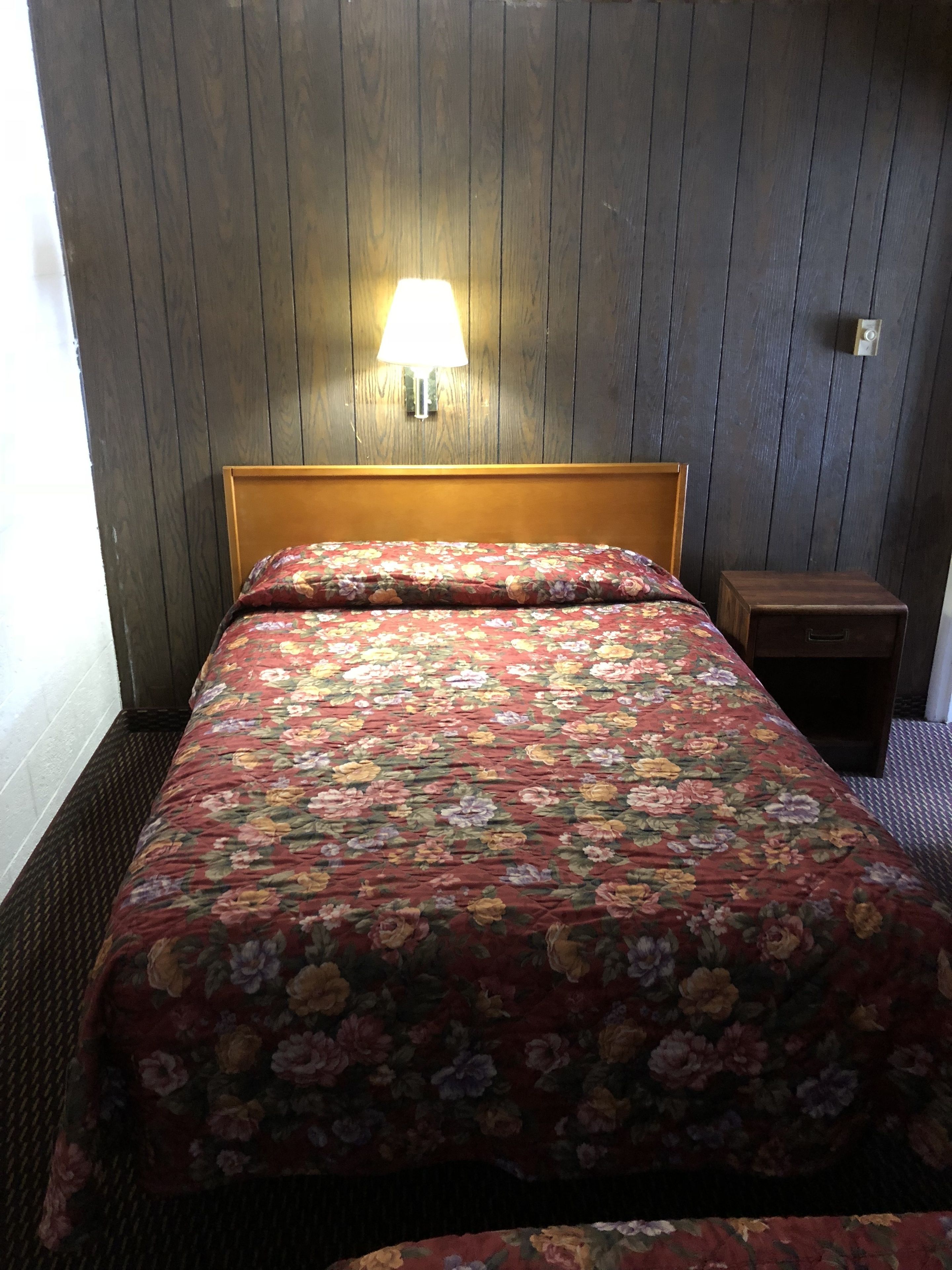 Une chambre avec une tête de lit en bois, une couvre-lit à motifs floraux, une table de chevet et une lampe fixée au mur.