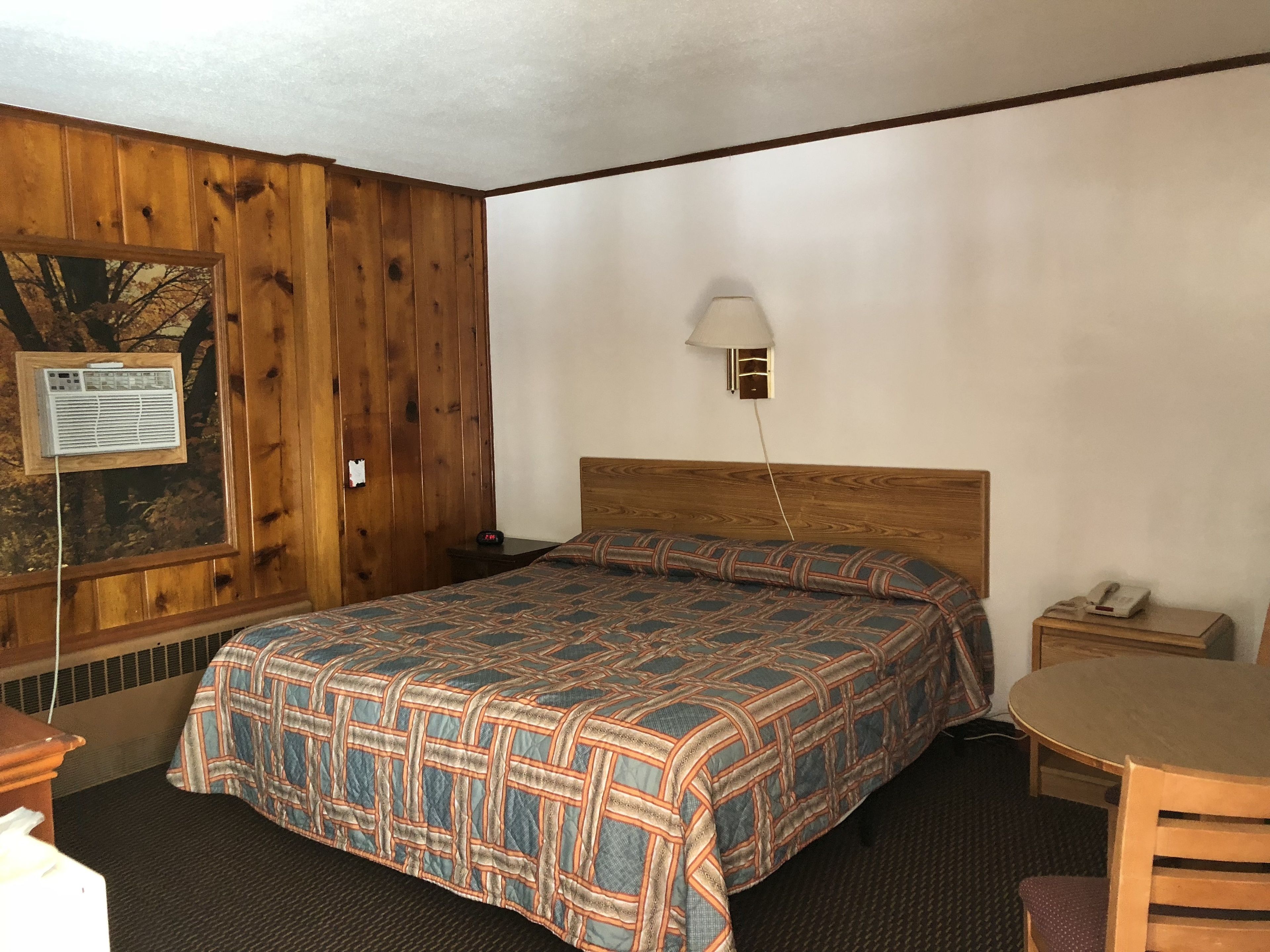 A hotel room with a wooden paneled wall, a bed with a patterned bedspread, a bedside table with a lamp, a small round table with a chair, and a wall-mounted telephone.