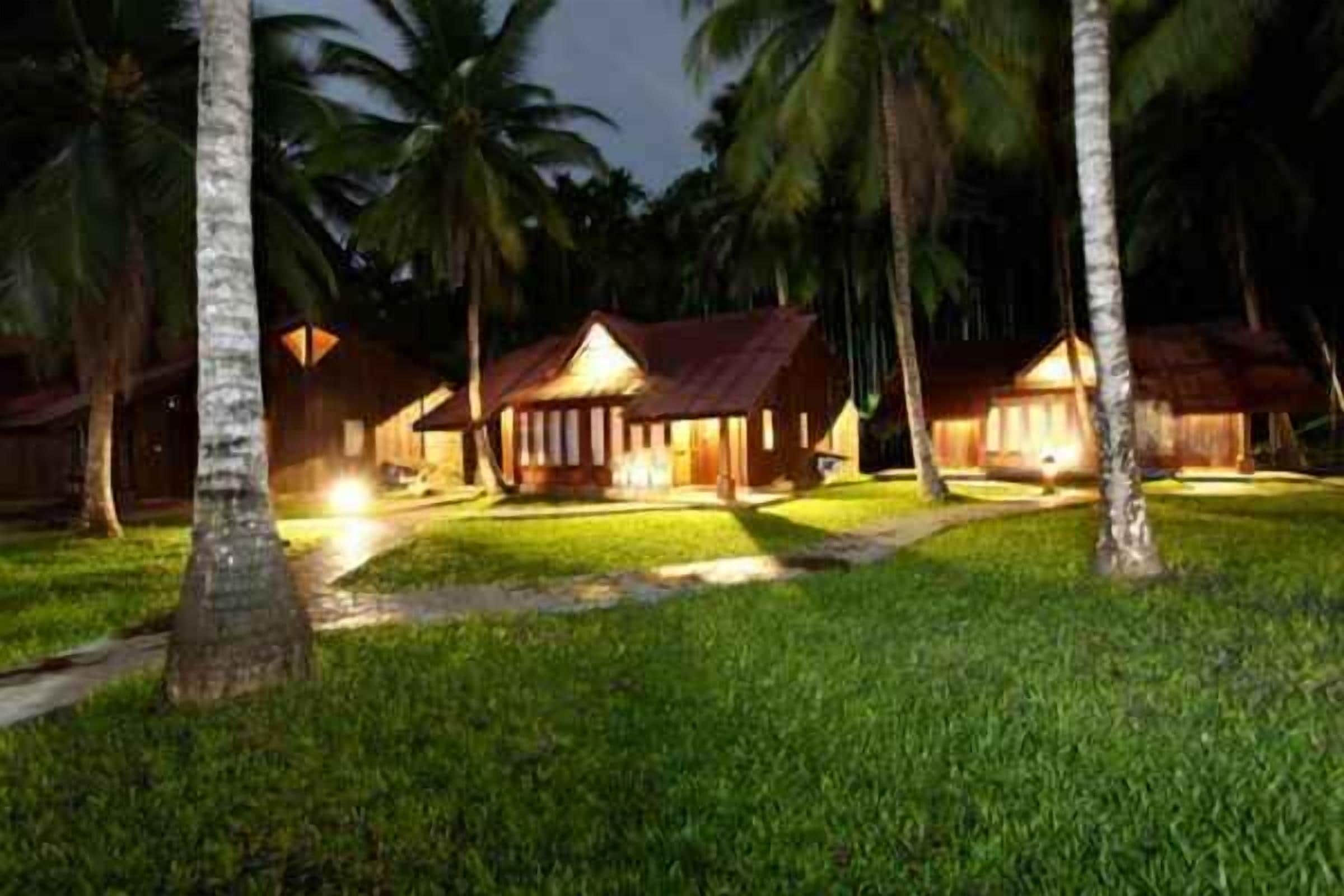 A row of wooden cottages with thatched roofs, surrounded by well-maintained grass and palm trees, indicating a tropical resort setting.