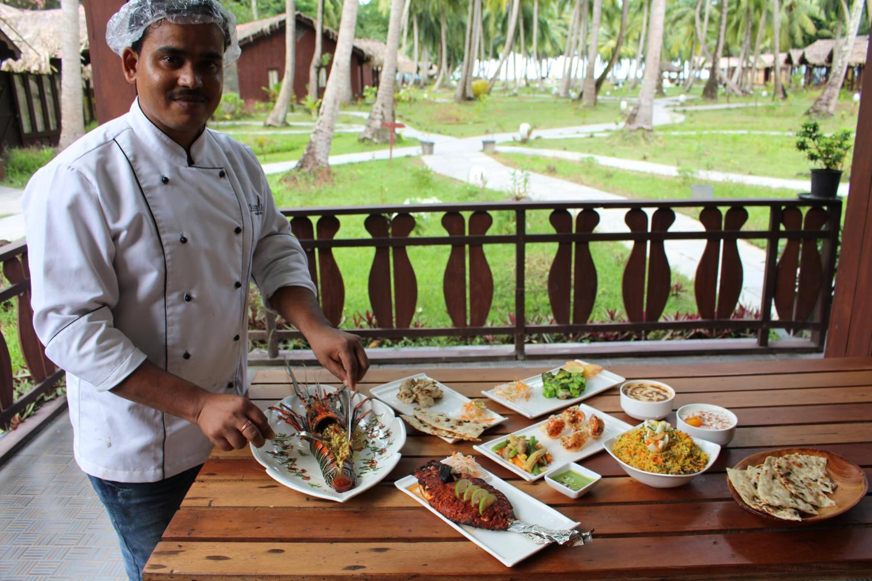A chef serving food on a wooden table with various dishes, set against a backdrop of a garden with palm trees.