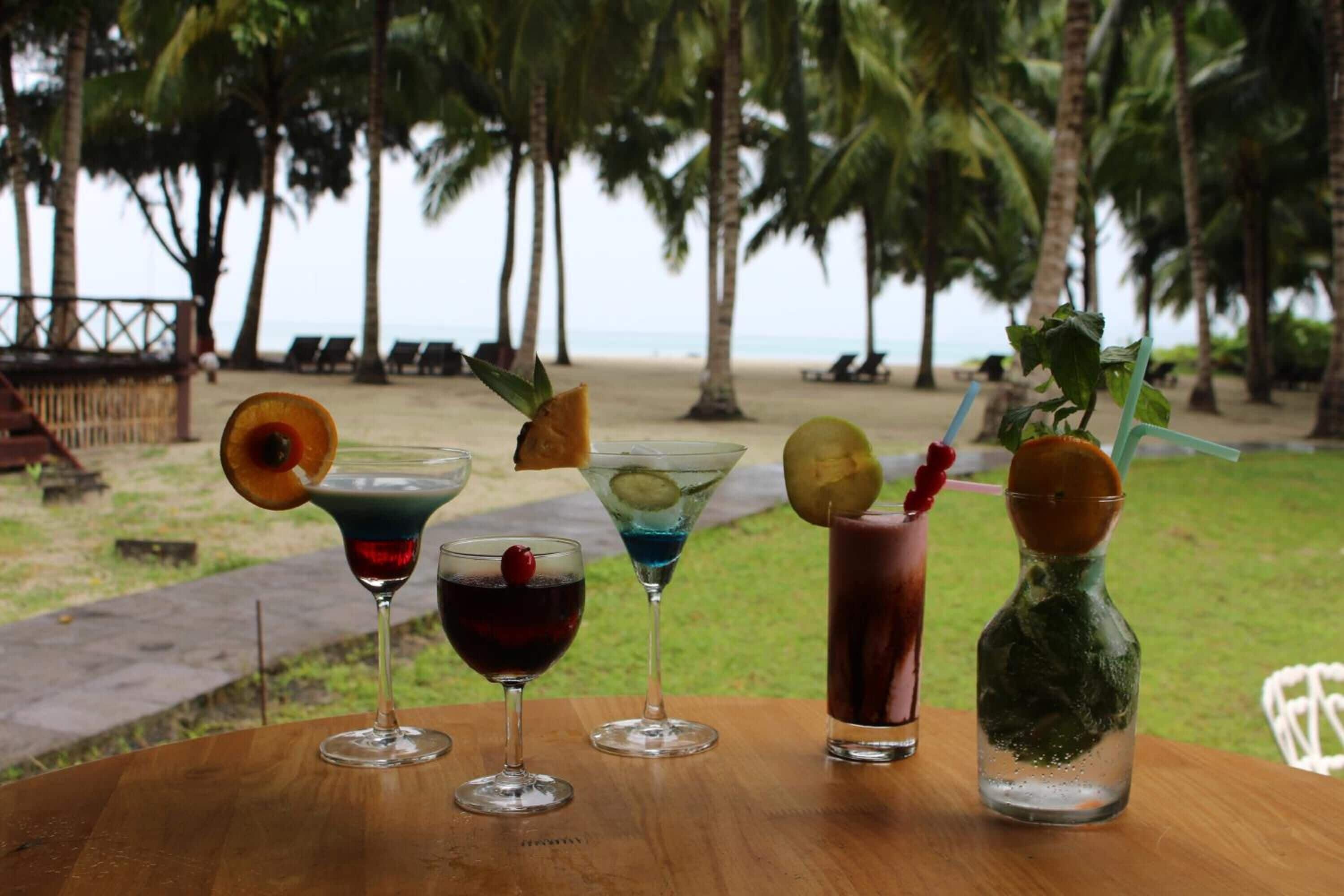 Una mesa de madera con cuatro bebidas, cada una con una guarnición diferente, en una playa con palmeras y una valla de fondo.
