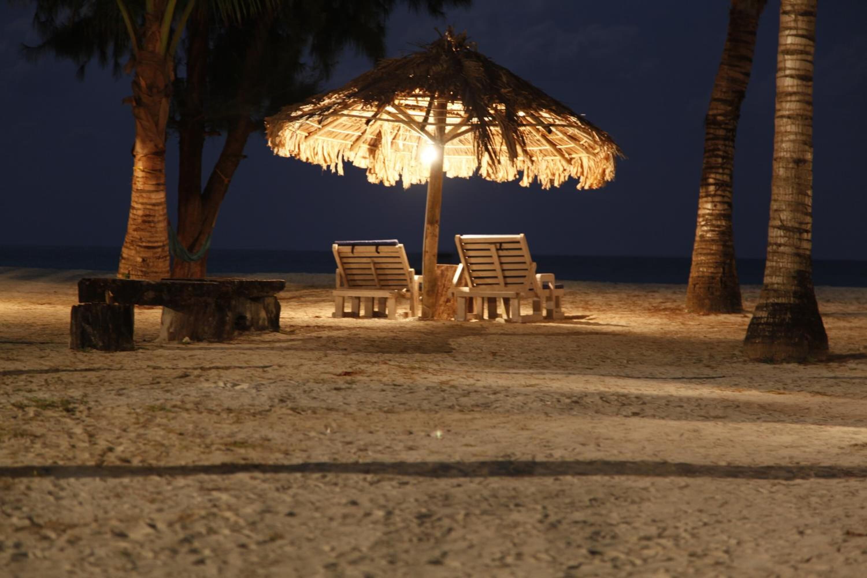 A beach scene with a thatched umbrella, two wooden chairs, and a bench.