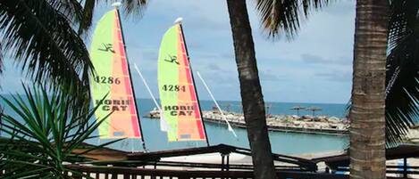 Vista sulla spiaggia con due barche a vela, palme e una recinzione in legno.