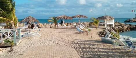 A beach with a paved walkway leading to the sea, lounge chairs, umbrellas, and palm trees.