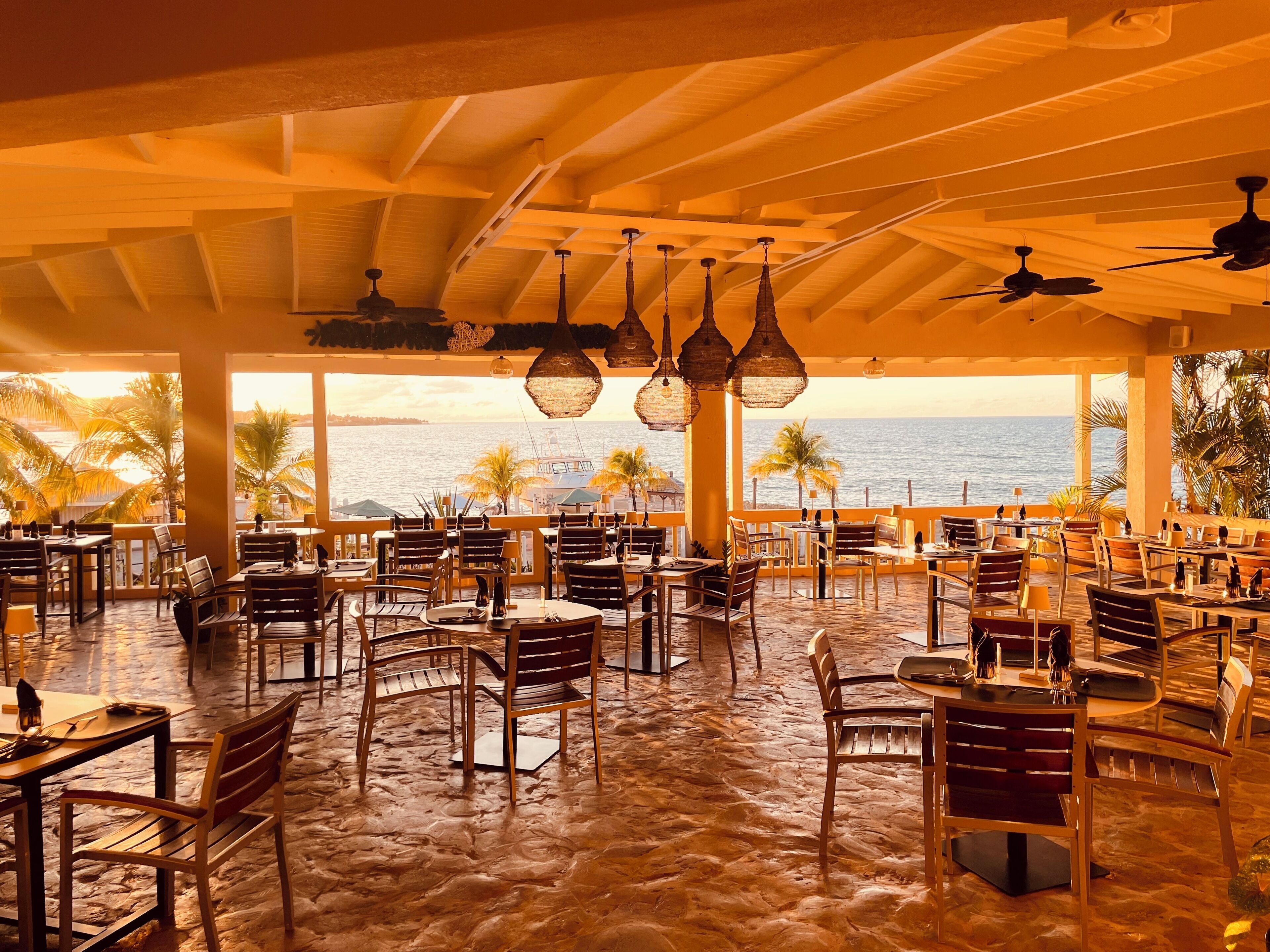 A restaurant with outdoor seating overlooking the sea, featuring wooden tables and chairs, hanging lights, and ceiling fans.