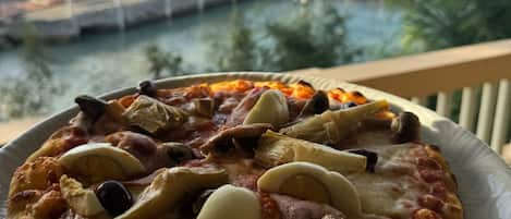 Pizza met diverse toppings op een bord, voor een achtergrond van een kustgezicht met palmbomen en een steiger.