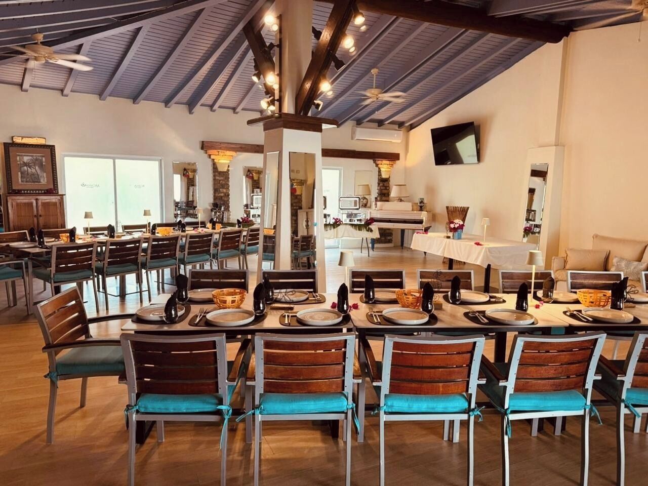 A spacious dining area with multiple tables and chairs, a high ceiling with exposed beams, and a television mounted on the wall.