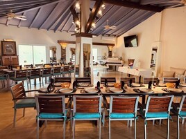 A spacious dining area with multiple tables and chairs, a high ceiling with exposed beams, and a television mounted on the wall.