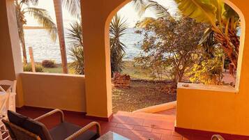 A room with a view of the sea, a wicker chair, a small table, and a window with a view of palm trees and a banana plant.
