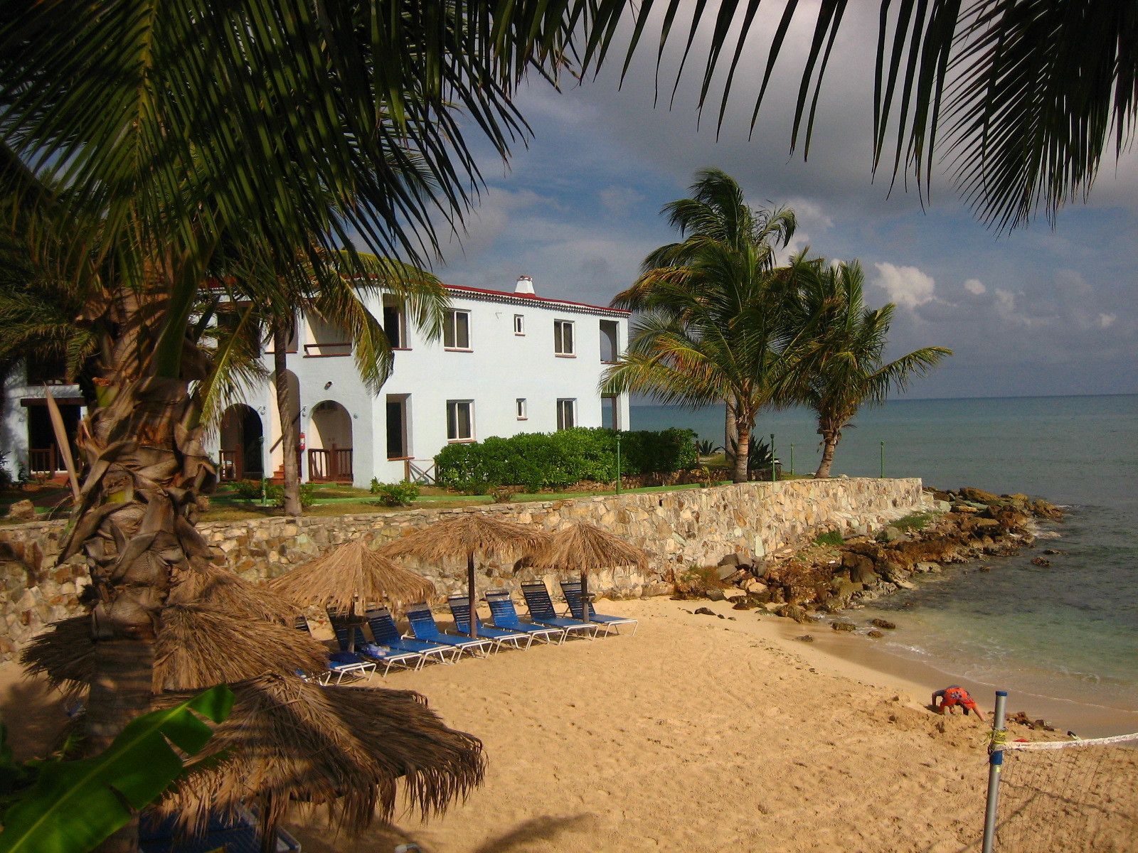A beachfront property with a white building, palm trees, and a sandy beach with lounge chairs.