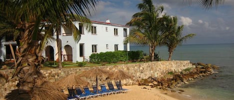 A beachfront property with a white building, palm trees, and a sandy beach with lounge chairs.