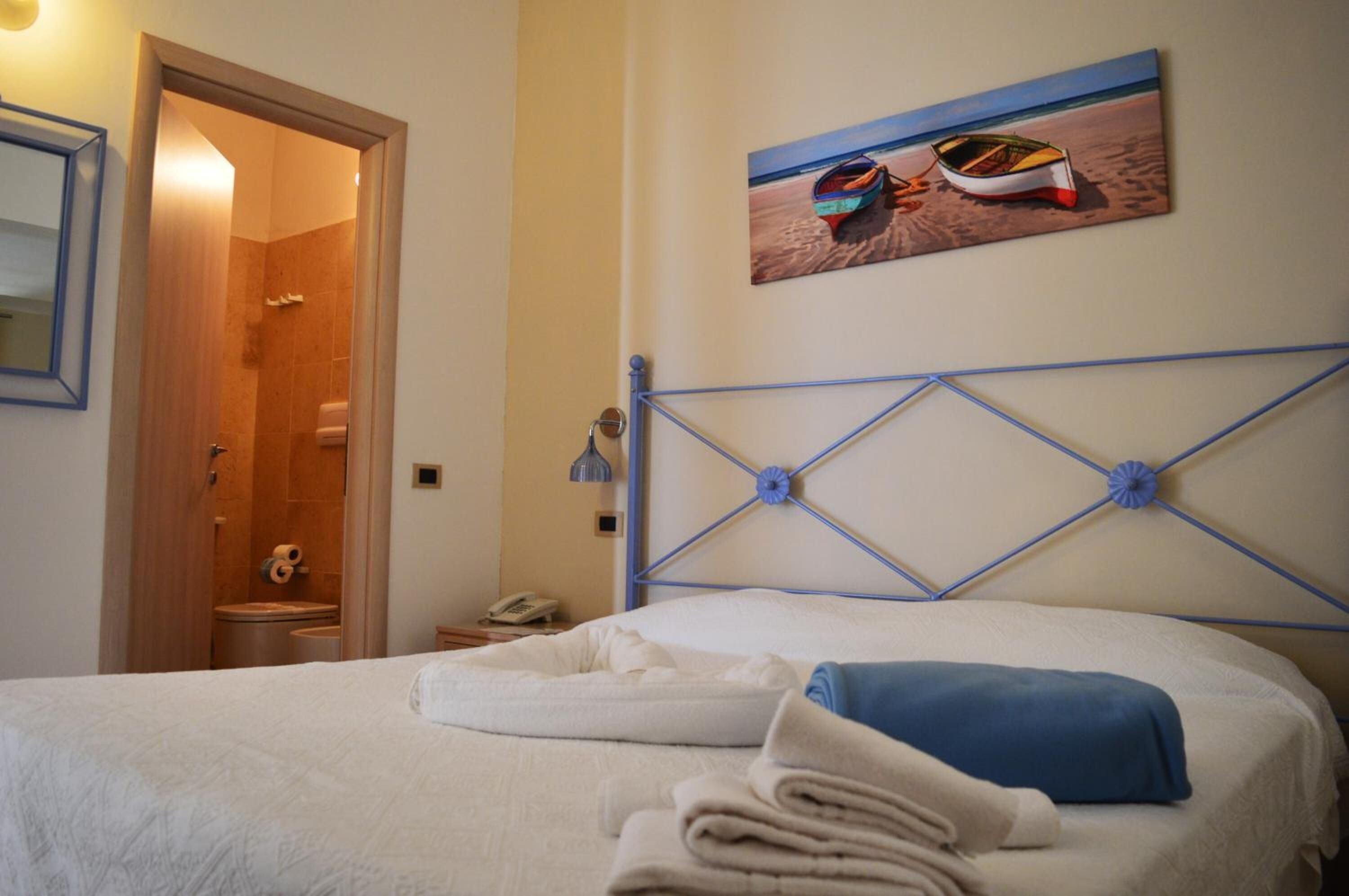 A neatly made bed with folded towels, a painting of boats on the wall, and a bathroom visible through an open door.