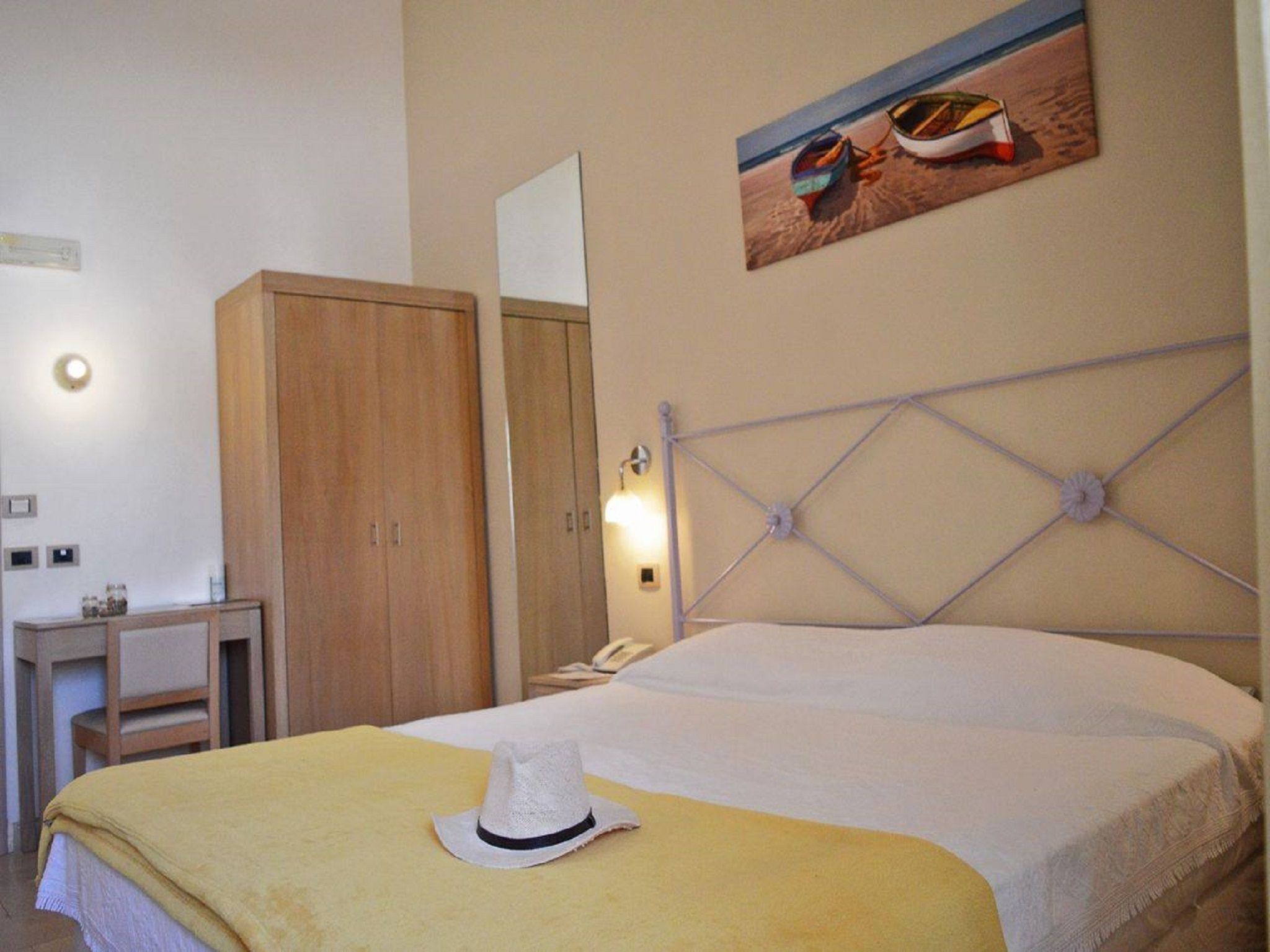 A hotel room with a bed, a wardrobe, a desk, and a painting of boats on the wall.