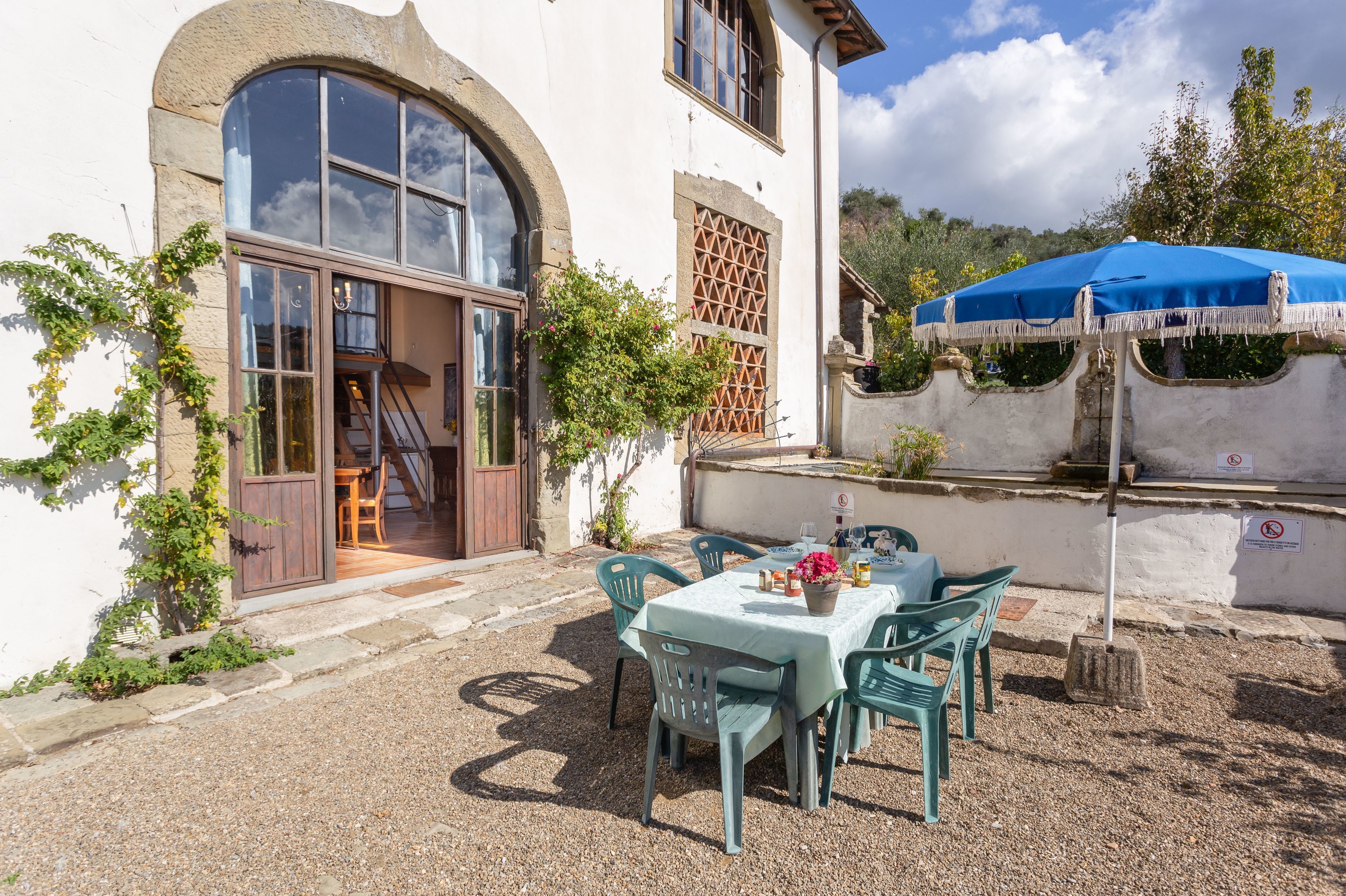 A patio with a table and chairs, a blue umbrella, and a building with a large arched window and a door.