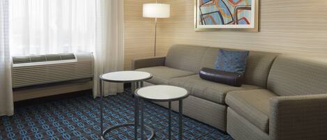 Habitación de hotel con sofá, dos mesas redondas, una lámpara, un cuadro abstracto enmarcado y una alfombra estampada.