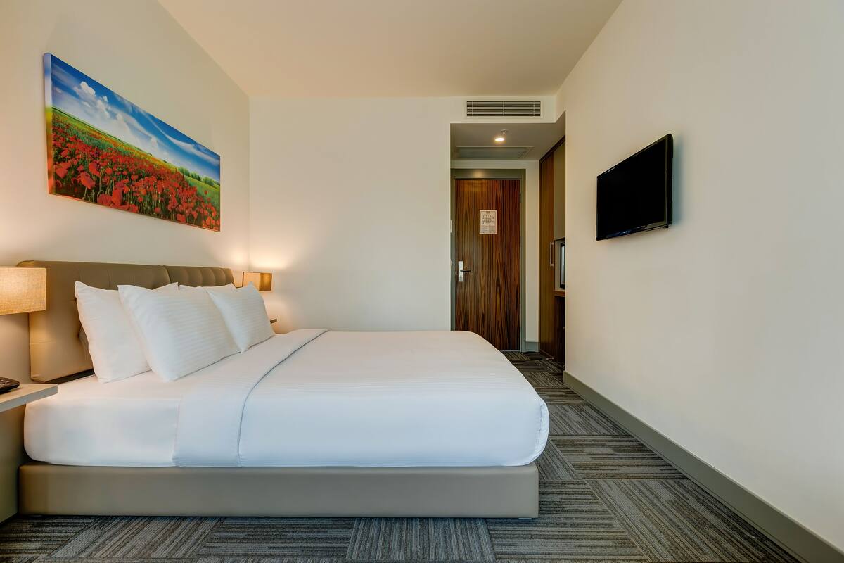 Une chambre d’hôtel avec un grand lit, un téléviseur à écran plat, un tableau représentant un champ et un sol recouvert de moquette.