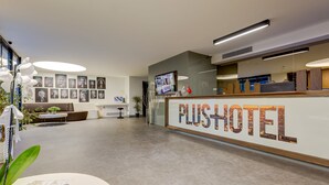 Ein moderner Hotel-Empfangsbereich mit einem 'PLUS HOTEL'-Schild, einem Empfangstresen, einer Sitzecke und gerahmten Bildern an der Wand.
