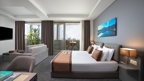 Ein modernes Hotelzimmer mit einem großen Bett, einer Couch, einem Schreibtisch und einem Fernseher. Ein Fenster bietet Blick auf die Stadt, und an der Wand hängt ein Gemälde.