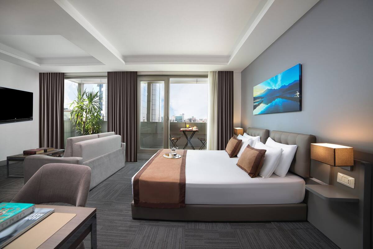 Une chambre d’hôtel moderne avec un grand lit, un canapé, un bureau et une télévision. Il y a une fenêtre donnant sur la ville et un tableau accroché au mur.
