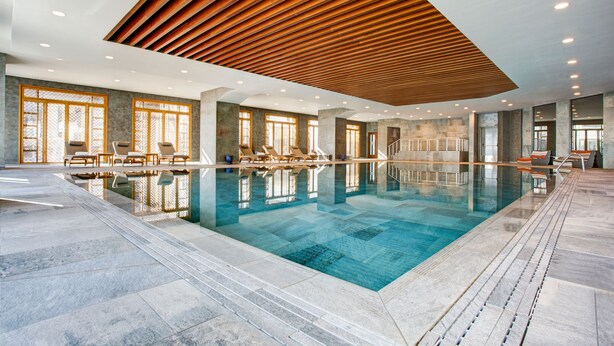 Une grande piscine intérieure à l’eau claire, avec un plafond en lambris de bois et des chaises longues disposées le long des bords.
