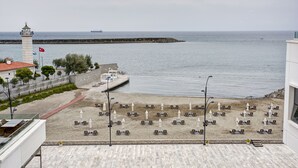 Une plage bordée de rangées de chaises longues et de parasols, un phare et un navire à l’horizon.