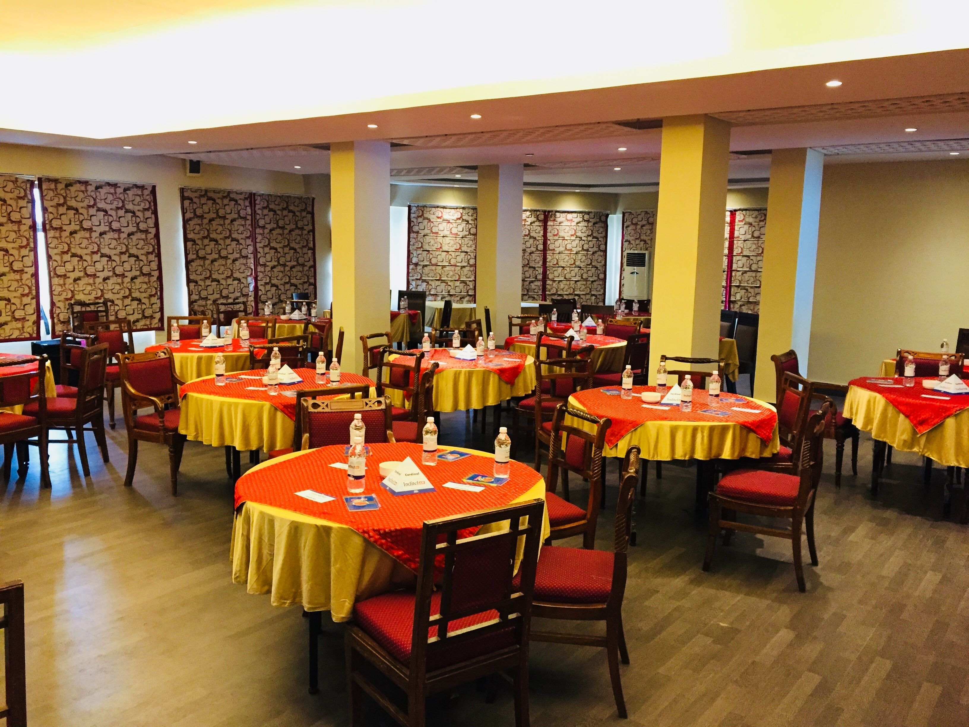 Un'ampia sala da pranzo con diversi tavoli rotondi ricoperti da tovaglie rosse, sedie in legno e una parete decorata sullo sfondo.