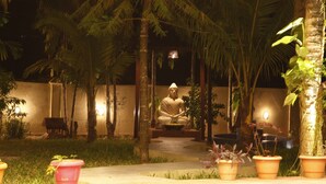 Una estatua de una figura sentada en un jardín con plantas en macetas y palmeras.