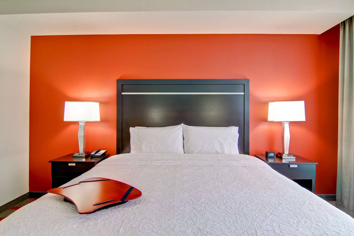 A hotel room with a large bed, two bedside tables with lamps, a nightstand, and a red pillow.