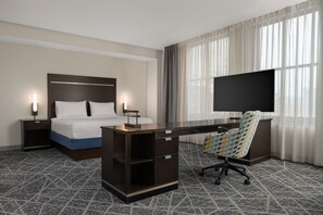Habitación de hotel con cama, escritorio, silla, televisor y cortinas.