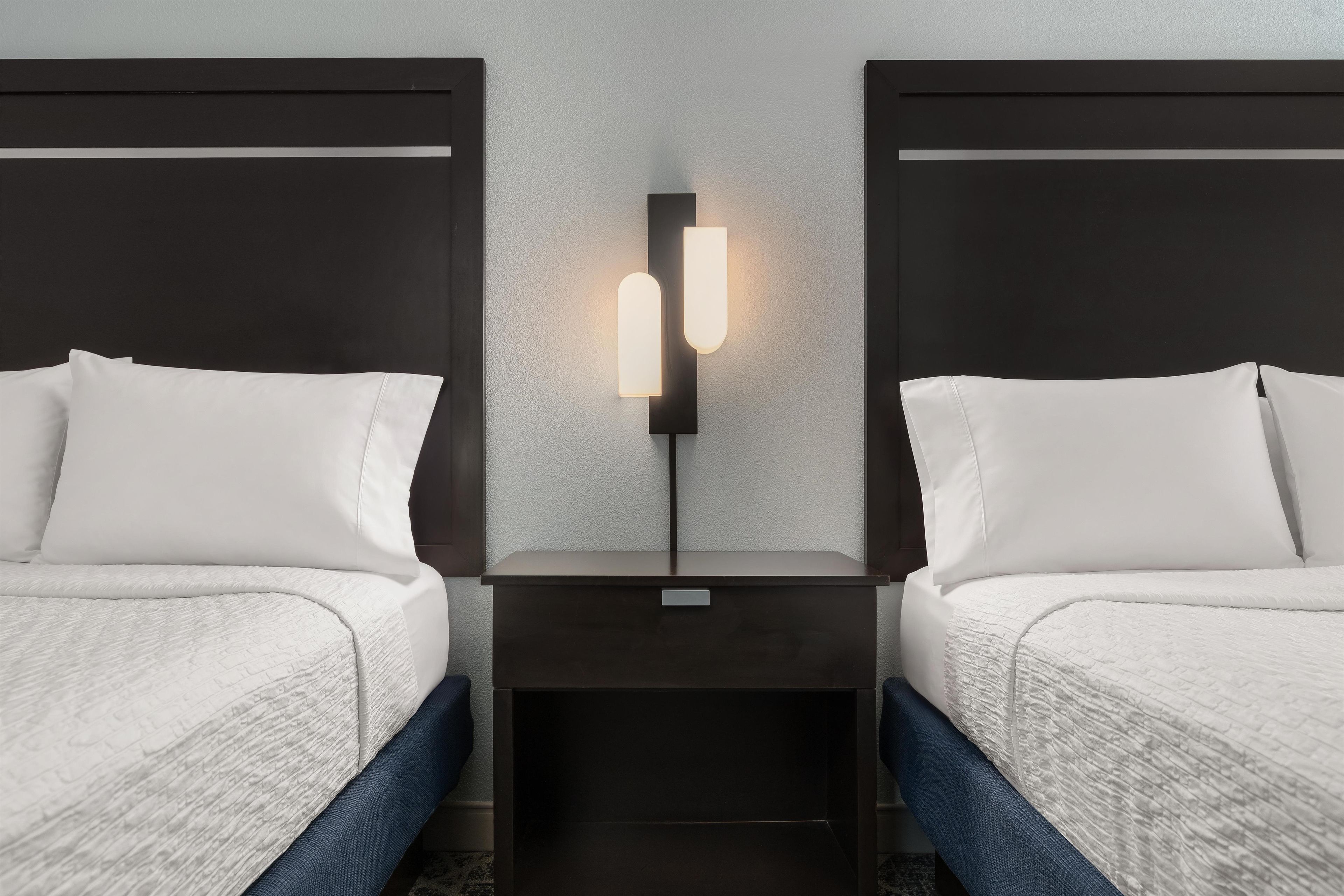 A hotel room with two beds, a nightstand, and a wall-mounted light fixture.