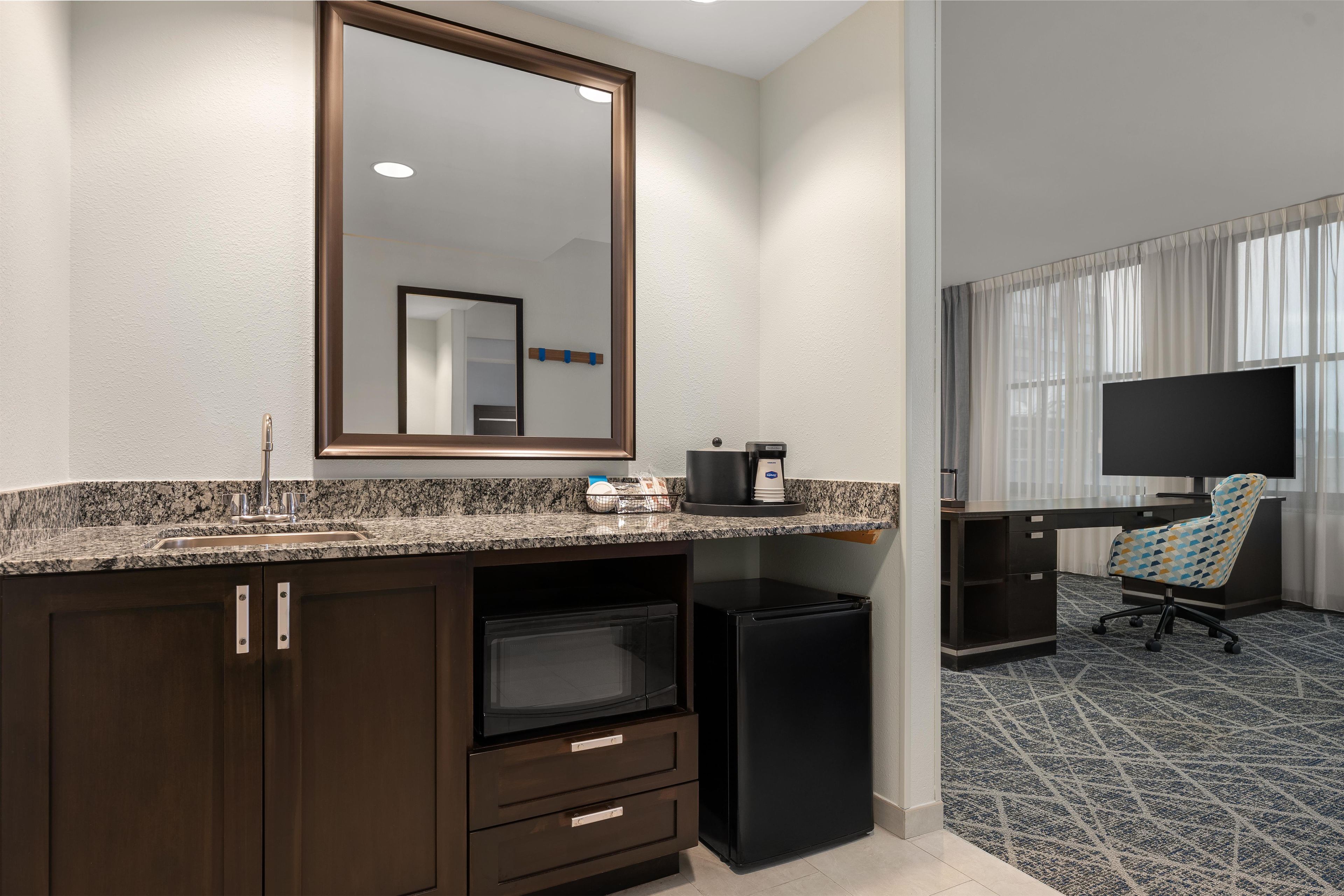 Habitación de hotel con encimera de granito, cafetera, refrigerador, escritorio con silla y televisor.