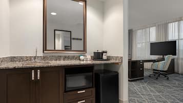 Habitación de hotel con encimera de granito, cafetera, refrigerador, escritorio con silla y televisor.