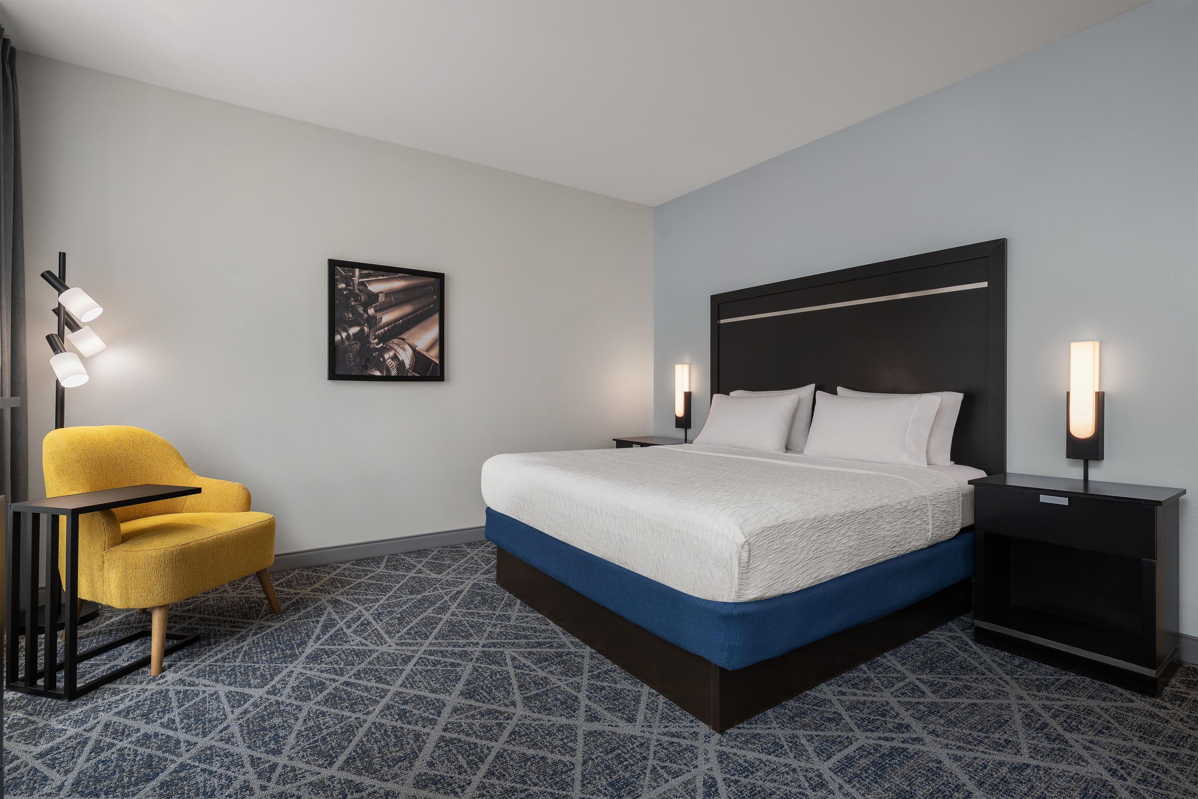 A hotel room with a bed, a chair, a nightstand, and a wall-mounted picture.