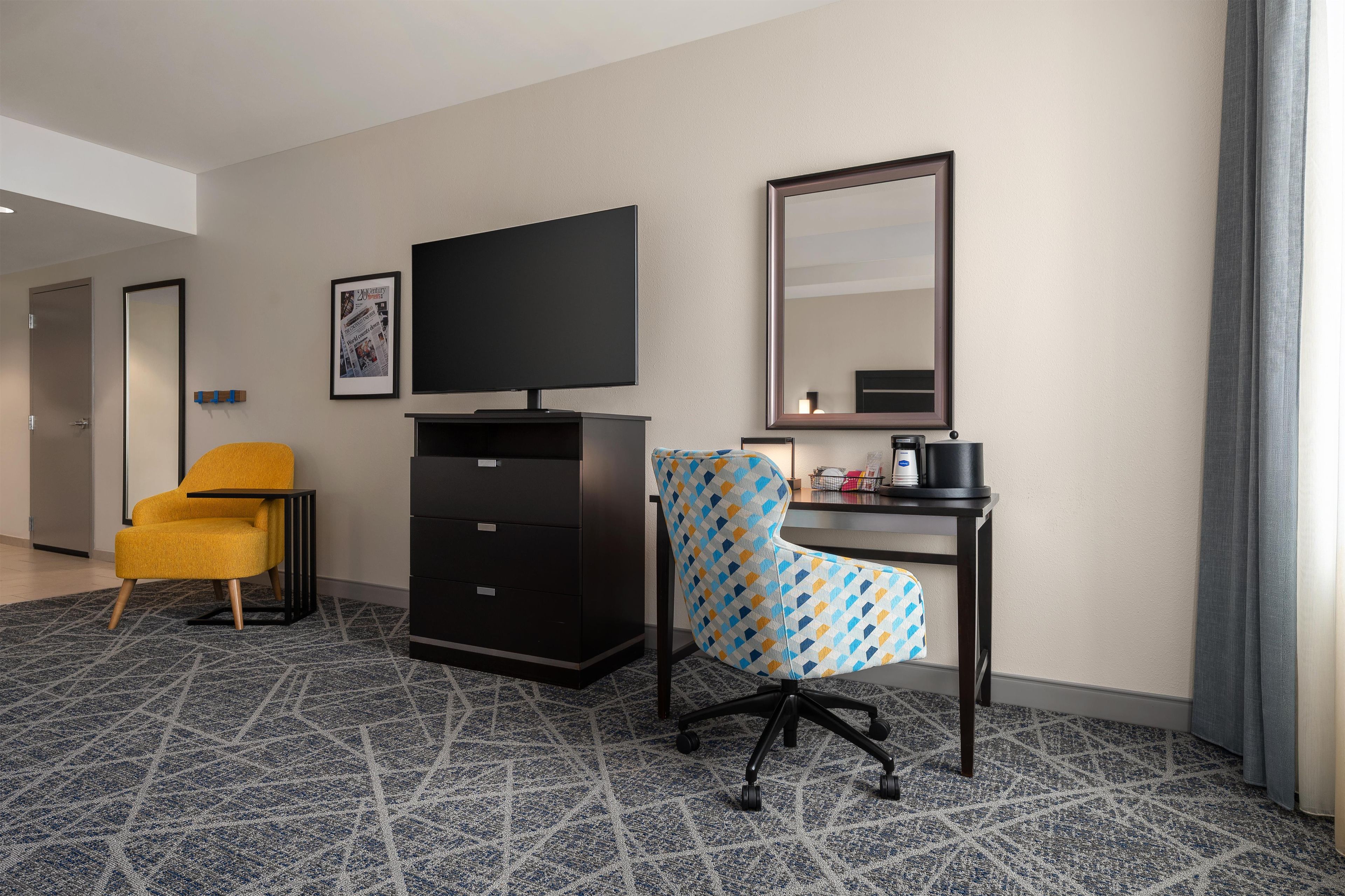 A hotel room with a flat-screen TV, a desk with a coffee maker, a chair, and a patterned carpet.