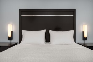 Una cama con cabecera oscura, dos almohadas blancas y dos lámparas de noche con forma rectangular y una fuente de luz en la parte inferior.