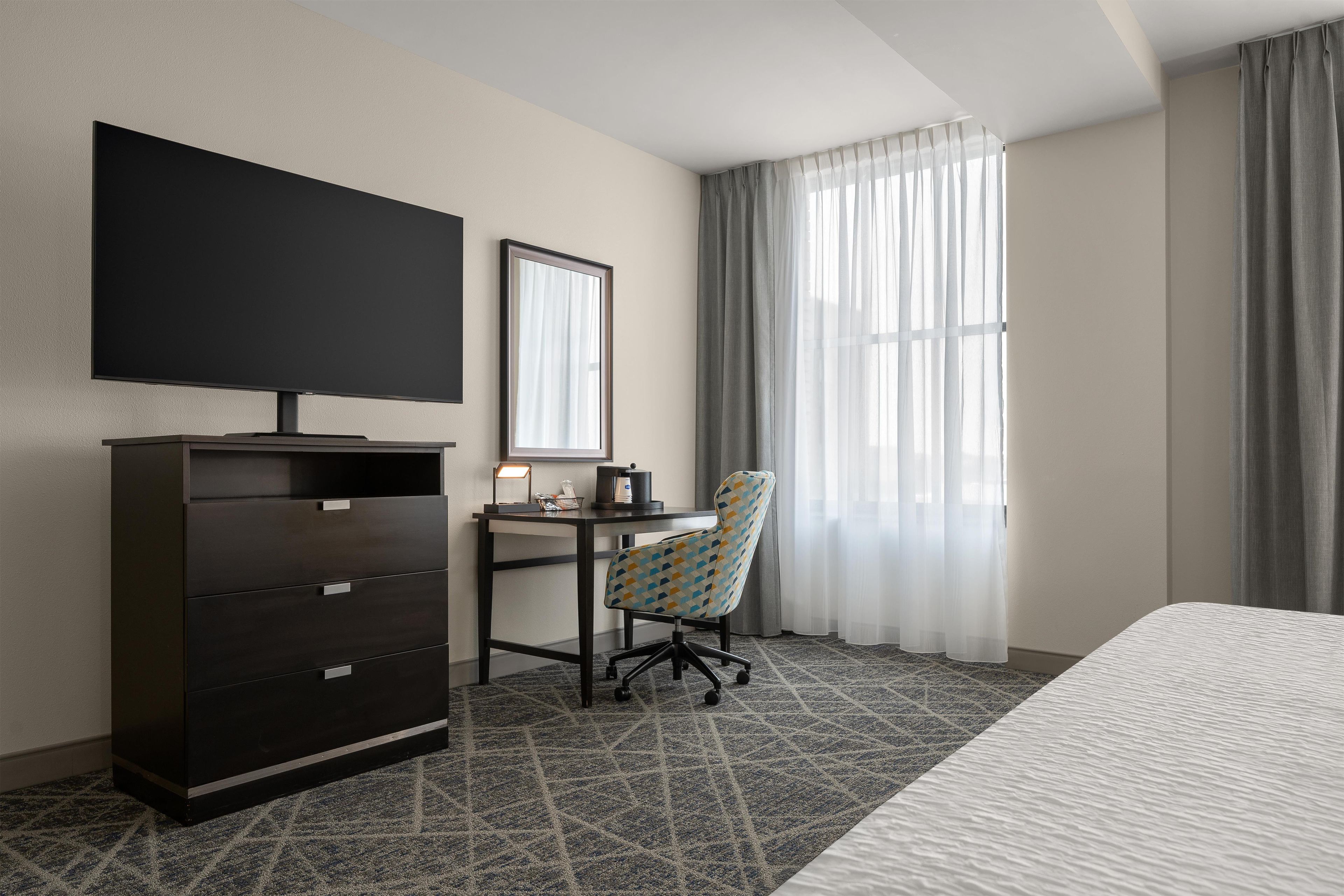 Habitación de hotel con cama, escritorio con silla, televisor, armario y ventana con cortinas.